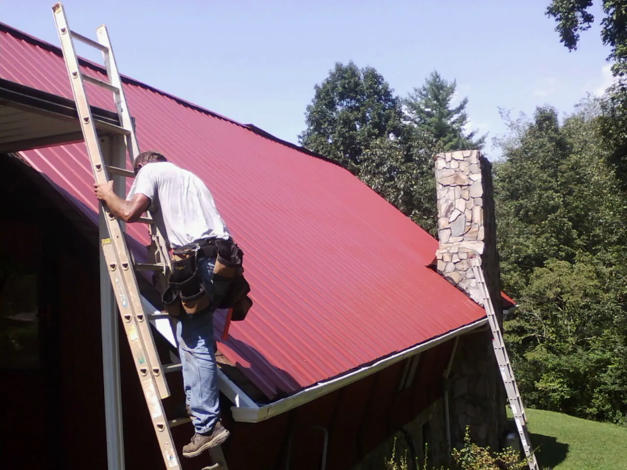Roofer on a ladder beside a red metal roof and stone chimney — Soffit & Fascia Repair by Roofing Brothers in Riverside